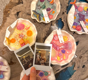 A hand holds two instant photos above several small decorative resin dishes containing colorful seashell candle and sea-themed embellishments, arranged on a rustic wood surface.