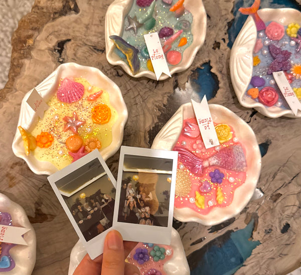 A hand holds two instant photos above several small decorative resin dishes containing colorful seashell candle and sea-themed embellishments, arranged on a rustic wood surface.