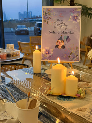 A custom printed birthday workshop game card titled 'birthday featuring a cartoon character and purple floral illustrations, displayed on a decorated table with lit candles and glassware.
