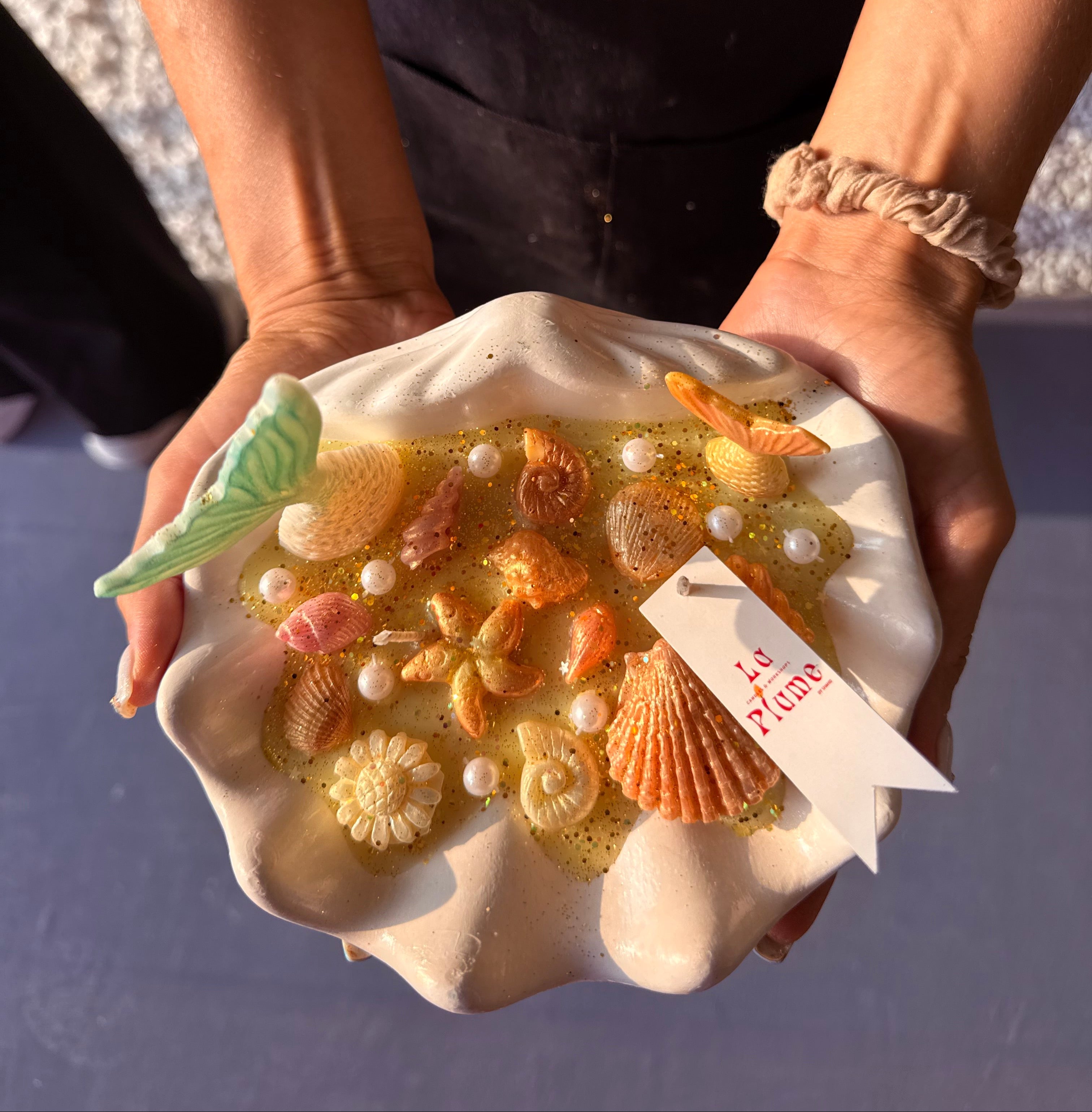 A person holds a decorative candle shell-shaped dish containing small seashell and coral-like embellishments in warm yellow, orange, pink, and green tones. The dish has a white scalloped rim and a small white tag attached with red text.
