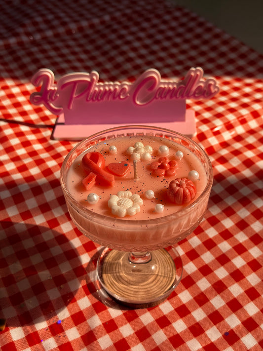A dessert-style candle in a clear glass bowl sits on a red-and-white checkered tablecloth. The candle wax is pink, topped with small white pearls, red wax accents, and decorative flower and ribbon shapes for breast cancer awareness month
